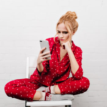 Serious girl in red sleepwear sitting with legs crossed on white background. Indoor portrait of upset young woman posing on chair with phone.